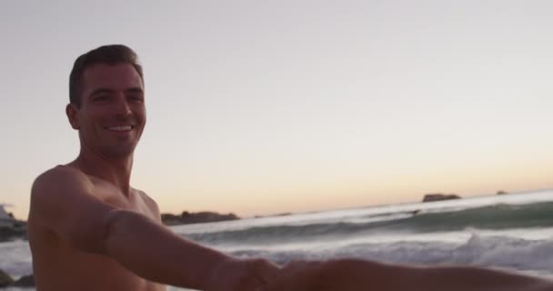 夕暮れ時に海でリラックスした幸せな白人青年のフロントビュー 彼の女性のパートナーの手を取り スローモーション — ストック動画