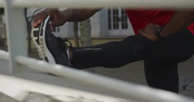 Spor kıyafetleri ve kulaklıklar takmış, ayakları duvara dayamış, şehirde antrenman yapmadan önce esneme hareketleri yapan, çitin arkasından yavaş çekim yapan Afro-Amerikan bir adamın ön görüntüsü.