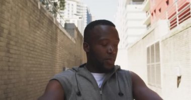 Spor kıyafetleri içinde, güneşli bir günde, sokakta esneyen ve kulaklık takan Afro-Amerikan bir adamın ön görüntüsü.
