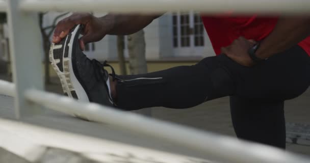 一名身穿运动服 头戴耳机 脚踏在墙上 在城市训练前伸展身体 通过栅栏 慢动作的黑人运动员的前视镜近在咫尺 — 图库视频影像