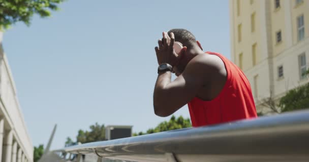 街を走る前にヘッドフォンをしてスポーツウェアを身に着けているアフリカ系アメリカ人の男性の側面図フェンスを通って見られるスローモーション — ストック動画