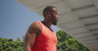 Spor kıyafetleri giymiş, güneşli bir günde şehirde antrenman yaparken akıllı bakışlarını kontrol eden Afro-Amerikan bir adamın ön görüntüsü, yavaş çekim.