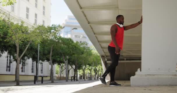 在一个阳光灿烂的城市里 一名身穿运动服的非裔美国人一边在训练 一边靠在柱子上 在桥下伸展着双腿 动作缓慢 — 图库视频影像