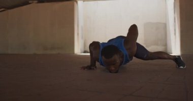Spor kıyafetleri içinde, güneşli bir günde şehirde antrenman yaparken gölgedeki köprünün altında silahlı şınav çeken Afrikalı Amerikalı bir adamın ön görüntüsü.