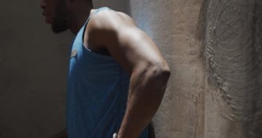 Spor kıyafetleri içinde, güneşli bir günde antrenman yaparken bir köprünün altından su içen ve duvara yaslanan Afro-Amerikan bir adamın yan görüntüsü.