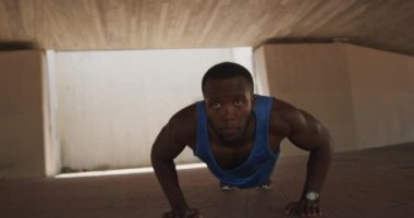 Spor kıyafetleri içinde, güneşli bir günde şehirde antrenman yaparken gölgede köprü altında şınav çeken Afro-Amerikan bir adamın ön görüntüsü.