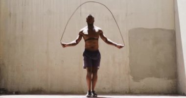 Göğüs kemeri takan, kalp atış hızı monitörü takan ve bir köprünün altındaki gölgede ip atlayan, güneşli bir günde egzersiz yapan, üstsüz bir Afrikalı Amerikalı adamın ön görüntüsü, yavaş çekim.