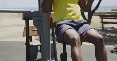 Spor kıyafetli Afrikalı Amerikalı bir adamın şehir parkında güneşli bir günde kulaklık takıp spor aletleriyle spor yaparken görüntüsü.