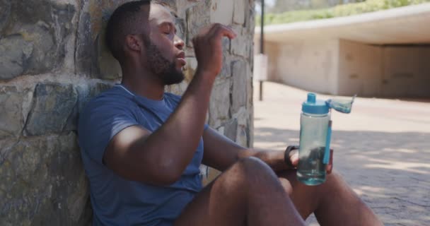 スポーツウェアを着たアフリカ系アメリカ人の男性が壁に座って 彼の額と飲料水をぬいぐるみながら 市内の晴れた日に訓練しながら ゆっくりとした動き — ストック動画