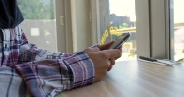 Kafenin penceresinin yanındaki masada oturan ve ağır çekimde akıllı telefon kullanan, karman çorman tişörtlü genç bir kadının yan görüntüsü.