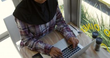 Kafedeki bir masada ağır çekimde tablet bilgisayar kullanan tesettürlü genç bir melez kadının yüksek açılı görüntüsü.