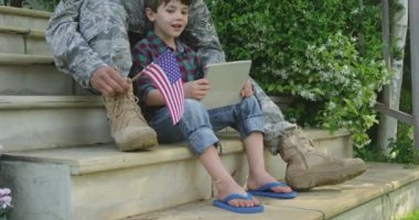 Evinin bahçesinde oğluyla merdivenlerde oturan, tablet bilgisayar kullanan ve elinde ABD bayrağı tutan, gülümseyen, ağır çekim gülümseyen yetişkin bir erkek askerin yan görüntüsü.