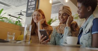 Türbanlı genç bir kadın, uzun kızıl saçlı beyaz bir kadın ve bir kafede oturup konuşan ve kulaklıklarını paylaşan genç bir Afro-Amerikan kadın birlikte akıllı telefondan müzik dinliyorlar.