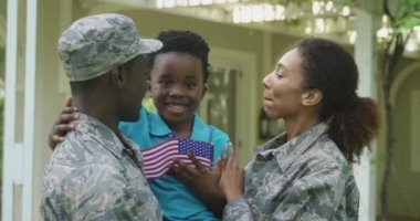 Genç, Afro-Amerikan bir erkek askerin ve melez bir kadın askerin evlerinin bahçesinde, gülümseyen ve ağır çekimde Amerikan bayrağını tutan genç oğullarını öperken ve kucaklarken ön görüntüsü.