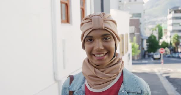 カメラに微笑む日当たりの良い街の通りに立ってヒジャーブを身に着けている若い混合人種の女性の肖像画 スローモーション — ストック動画