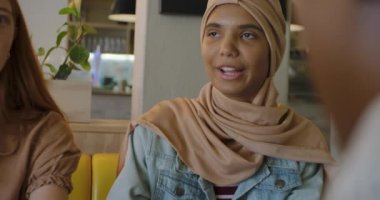 Ön planda, kafeteryada arkadaşlarıyla oturan, konuşan, gülen, yavaş çekim yapan, tesettürlü genç bir ırk kadını var.