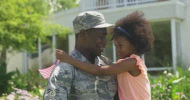 Side view close up of a young adult African American male soldier in the garden outside his home, holding and embracing his young daughter, who is holding a US flag, they are looking at each other smiling, slow motion