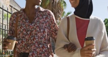 Afrika kökenli genç bir Amerikalı kadın ve tesettürlü genç bir melez kadın güneşli bir caddede kol kola yürüyorlar ve konuşuyorlar arkalarında ağaçlar ve kahveleri tutuyorlar.