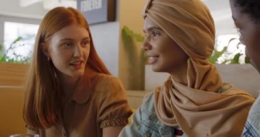 Ön planda, tesettürlü genç bir ırk kadını ve uzun kızıl saçlı beyaz bir kadını bir kafede bir arkadaşıyla oturup konuşurken ve gülerken görüyoruz.