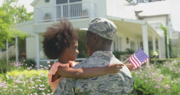 一位年轻的非裔美国士兵在自家门外的花园里 紧紧地抱住他的小女儿 紧紧地抱着他的后视镜 小女儿正对着摄像机 闭上眼睛微笑着 举着一面美国国旗 动作缓慢 — 图库视频影像