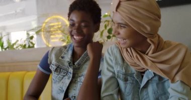 Ön planda genç bir Afro-Amerikalı kadın ve tesettürlü genç bir melez kadın birlikte bir kafede oturuyor, konuşuyor ve gülüyor, yavaş çekim.