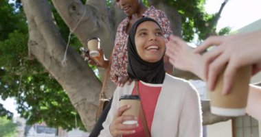 Yan görüntüde genç beyaz bir kadın, genç bir Afrikalı Amerikalı kadın ve tesettürlü genç bir ırk kadını güneşli bir şehir sokağında oturmuş, bir ağacın kenarında oturmuş, kahvelerini yudumluyor, birlikte konuşuyor ve gülüyorlar, yavaş çekim.
