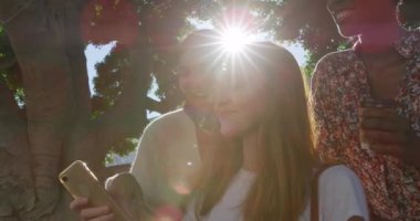 Ön cephede genç beyaz bir kadın, genç bir Afrikalı Amerikalı kadın ve genç bir melez kadın güneşli bir şehir sokağında ağaç kenarında dikilip birlikte akıllı telefona bakıyor ve gülüyorlar.