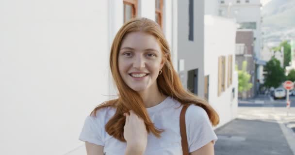 若い白人女性の肖像画の近くに長い赤い髪の日当たりの良い街の通りに立ってカメラに笑みを浮かべて スローモーション — ストック動画