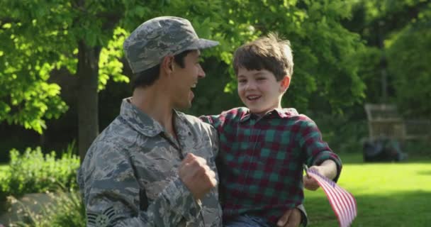 一个年轻的男女混血儿背着他的小儿子 举着一面美国国旗 面带微笑 慢吞吞地看着对方 正站在屋外的花园里 近视着一个年轻的男女混血儿 — 图库视频影像