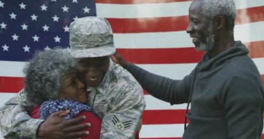 Genç, mutlu, Afro-Amerikan erkek askerin ön görüntüsü. Ailesiyle kucaklaşan, hepsi Amerikan bayrağı önünde gülümseyen, ağır çekimde.