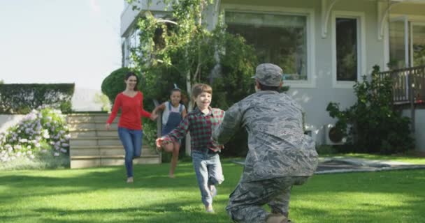 一个年轻的成年混血儿在自家门外的花园里跪着拾起他的小儿子 他的小儿子拿着一面美国国旗奔向他 他的母亲和姐姐手牵着手 在他身后奔跑 动作缓慢 — 图库视频影像