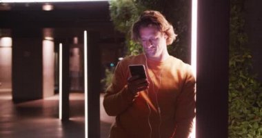 Kafkasyalı bir adam günlük kıyafetler ve kulaklıklar takıyor, gece boyunca şehir sokaklarında geziyor, direğe yaslanıyor ve akıllı telefonunu kullanıyor..