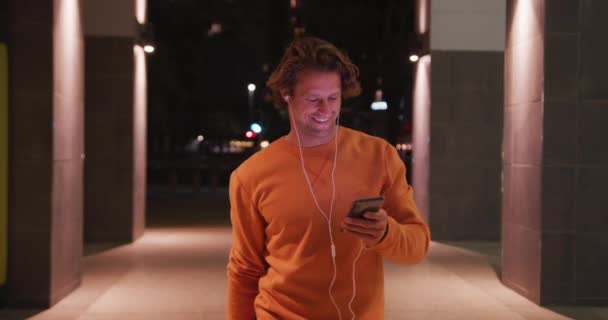 夜の街中で彼のスマートフォンと笑顔を使用して オレンジの引き出しやイヤフォンを身に着けている白人男性 — ストック動画