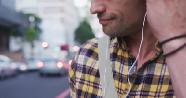 一名白种人 身穿格子衬衫 白天在城市街道上走来走去 用智能手机 戴上耳机听音乐 — 图库视频影像