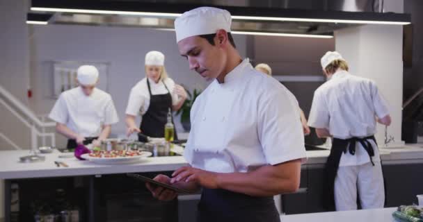 Portrét profesionálního smíšeného závodního šéfkuchaře v kuchařské uniformě v kuchyni restaurace, pomocí tabletu, při pohledu na kameru, s kolegy pracujícími v pozadí ve zpomaleném filmu