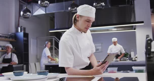 专业的白种人男性厨师在餐馆厨房里穿着白色厨师的衣服 用平板电脑 同事们在后台慢动作地工作 — 图库视频影像