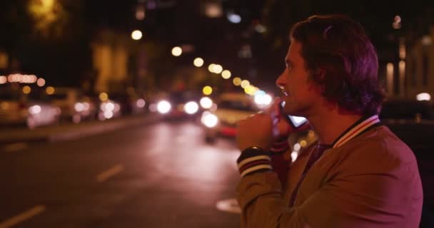 夜の街中でスマートフォンを片手に笑顔で話す白人男性 — ストック動画