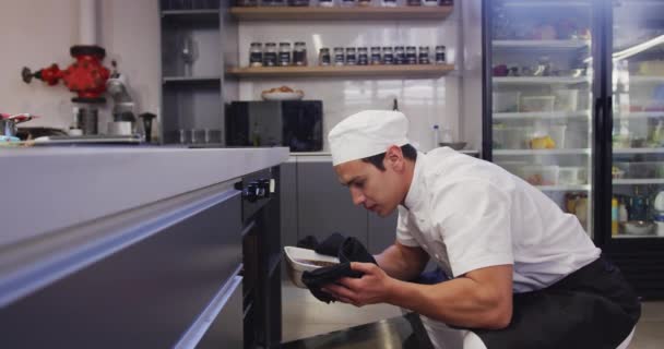 レストランのキッチンで白人シェフを身に着けているプロの混合レースの男性シェフは スローモーションでオーブンから食べ物を取ります — ストック動画
