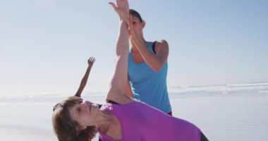 Çok ırklı bir grup bayan arkadaş güneşli bir günde kumsalda egzersiz yapmaktan zevk alıyor yoga yapıyor, beyaz bir bayan koçla yoga pozisyonunda esneme hareketleri yapıyor.