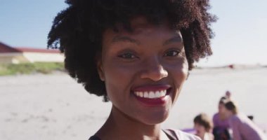Güneşli bir günde kumsalda egzersiz yapmaktan zevk alan, yoga yapan, kameraya bakan Afrikalı Amerikalı çekici bir kadının portresi.