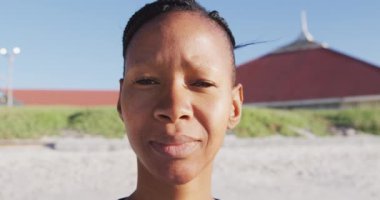 Güneşli bir günde kumsalda egzersiz yapmaktan zevk alan, yoga yapan, kameraya bakan melez bir kadının portresi.