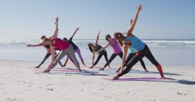 Çok ırklı bir grup kız arkadaş güneşli bir günde kumsalda egzersiz yapıyor yoga yapıyor, yoga pozisyonunda geriniyor..