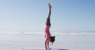Karışık ırk çekici bir kadın, spor kıyafetleri giyiyor, yoga yapıyor, yoga pozisyonunda, güneşli sahilde ayakta duruyor.