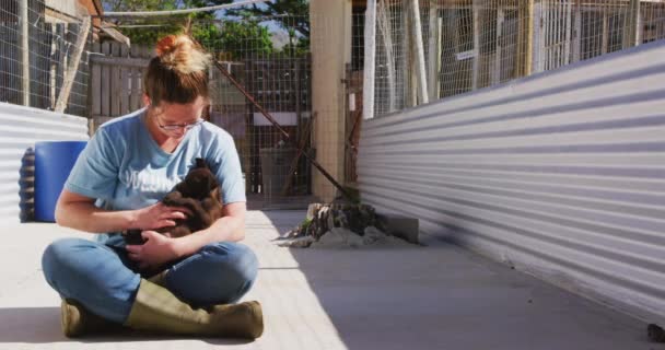動物の避難所で青い制服を着た白人女性のボランティア 彼女の腕の中で救助された子犬を座って保持し 遅い動きで晴れた日にカメラを見て笑顔 — ストック動画