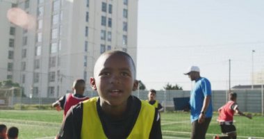 Güneşli bir günde sahada durup antrenman sırasında dinlenen ve arka planda takım arkadaşları olan Afrikalı Amerikalı bir futbolcunun portresi.
