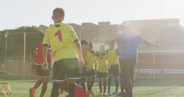 Çok ırklı erkek futbol oyuncularından oluşan bir grup güneşin altında bir oyun sahasında birbirlerine sarılıyorlar.