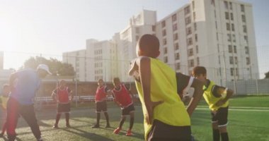 Karışık ırk erkek futbol koçu ayakta duruyor ve çok ırklı futbol oyuncularından oluşan bir gruba futbol antrenmanı sırasında güneşin altında esneme egzersizleri yaptırıyor.