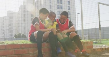 Takım elbiselerini giyen, oyun sahasının kenarında oturan, akıllı telefonları bir arada tutan ve kullanan çok ırklı bir grup genç futbolcu.