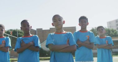 Çoklu etnik gruptan futbol oyuncuları takım elbiselerini giyiyor, güneşin altında bir oyun sahasında duruyor ve kollarını kavuşturup kameraya bakıyorlar.