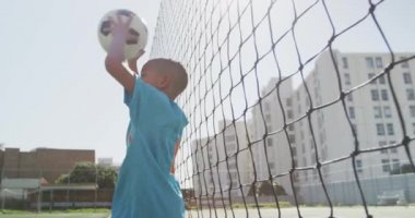 Güneşli bir günde, yeşil bir sahada futbol oynayan karışık yarışçı çocuk elinde bir top tutuyor ve fırlatıyor.